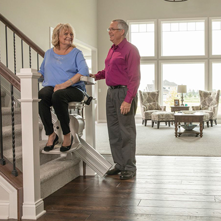 Woman riding Bruno Elan home stair lift while husband stands on bottom landing