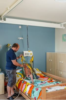 man using portable ceiling lift to safely transfer his kid from his bed to wheelchair