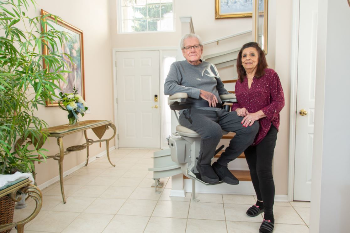 Lifeway Mobility stair lift customer sitting on his new stair lift as wife stands next to him at bottom of stairs